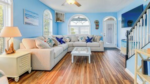 Ein Wohnzimmer mit einem Ecksofa, einem Couchtisch, Holzboden, einem Deckenventilator und einer Treppe mit weißen Geländern.