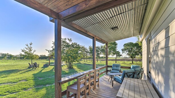 A covered outdoor deck with wooden furniture, a view of a grassy field, and a building in the background.