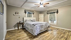 A bedroom with a wooden floor, a bed with a patterned bedspread, a ceiling fan, a window with curtains, and a small table with a lamp.