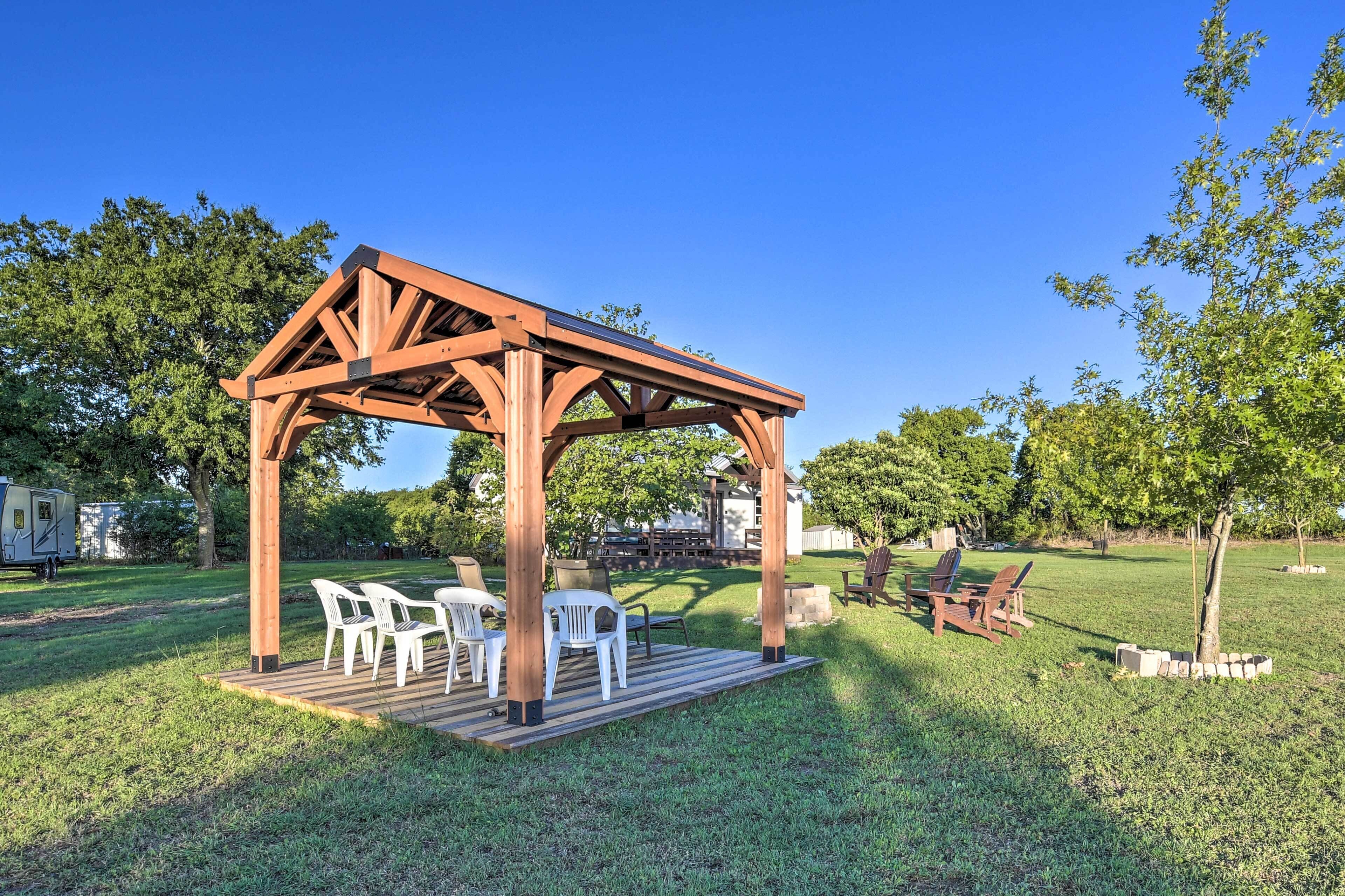A wooden pergola with white plastic chairs and a table, surrounded by a grassy yard and trees.