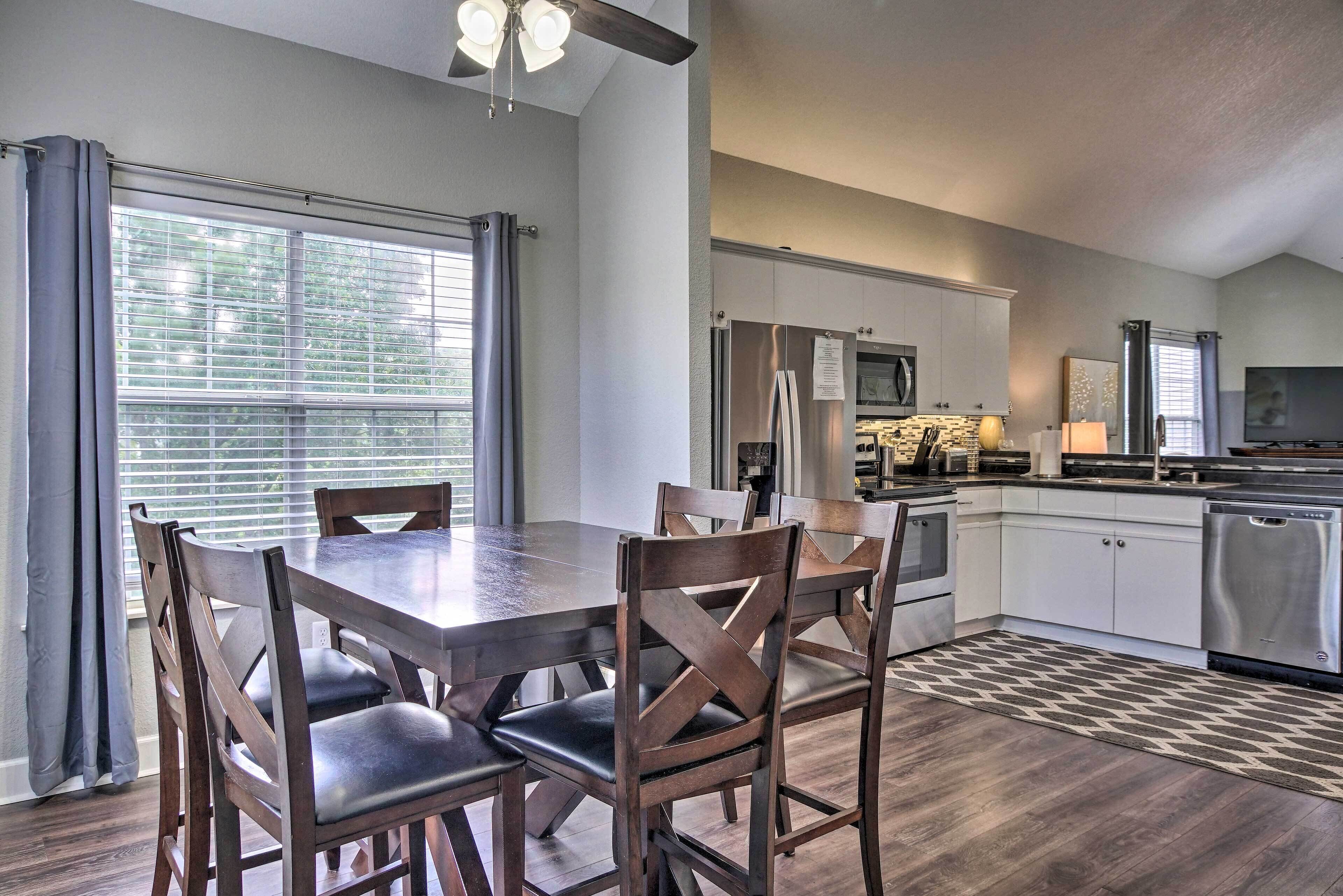 A dining area with a wooden table and chairs, a kitchen with modern appliances, and a ceiling fan.