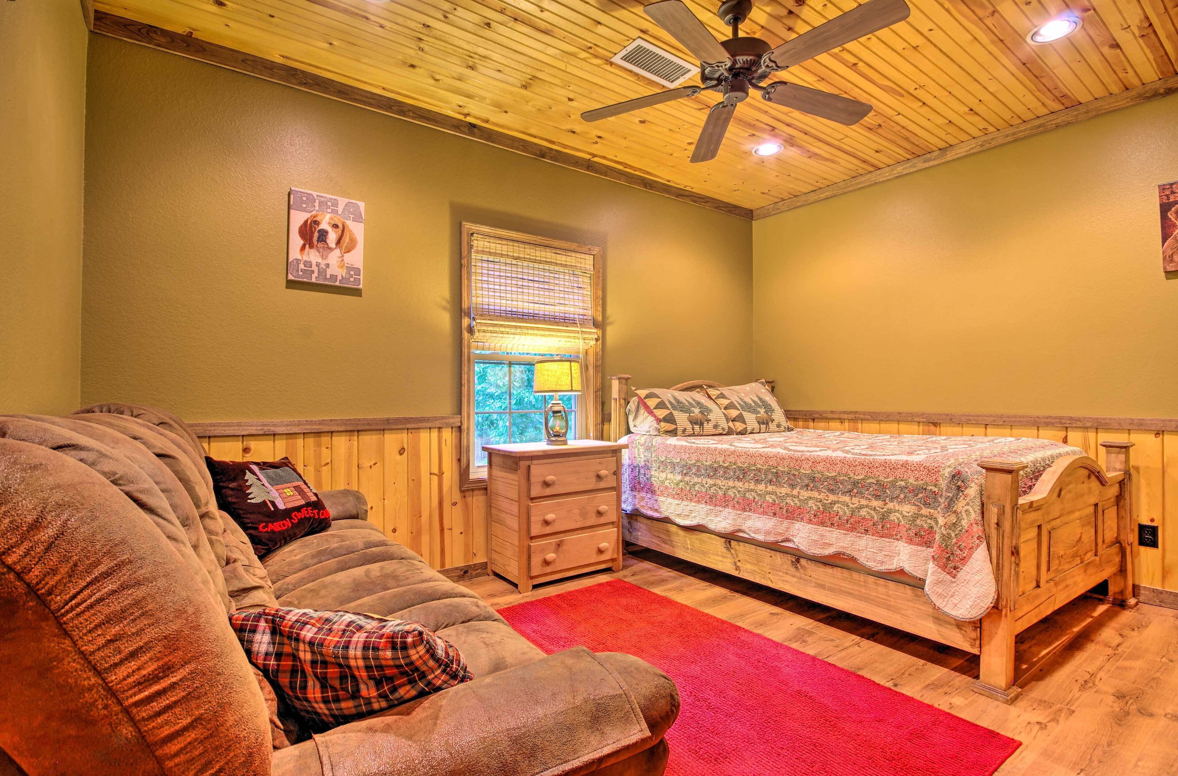A cozy bedroom with a wooden ceiling, a bed with a patterned bedspread, a wooden dresser, a red rug, and a couch with plaid cushions.