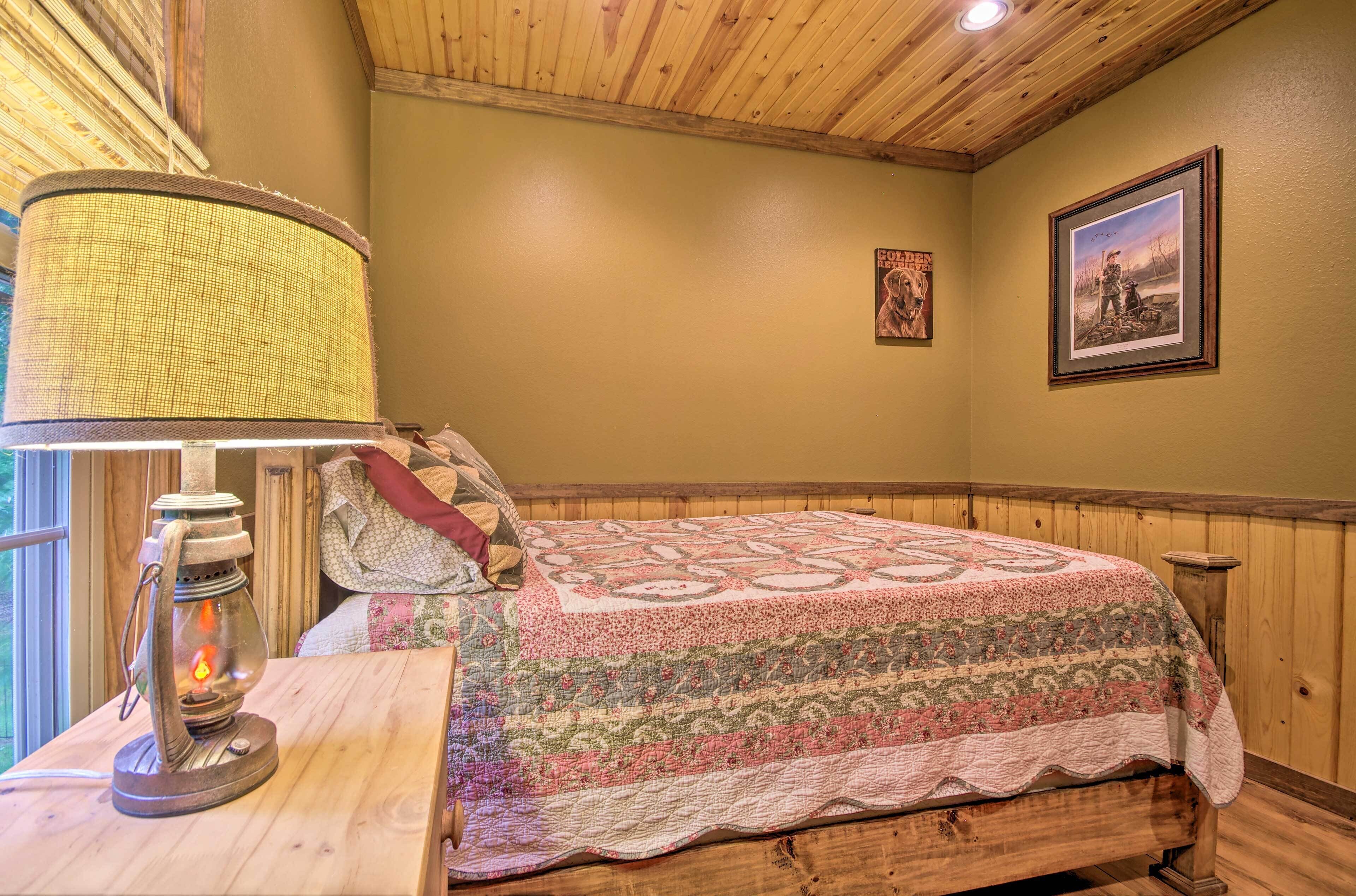 A cozy room with a wooden ceiling, a bed with a patterned quilt, a table with a lantern, and framed pictures on the wall.