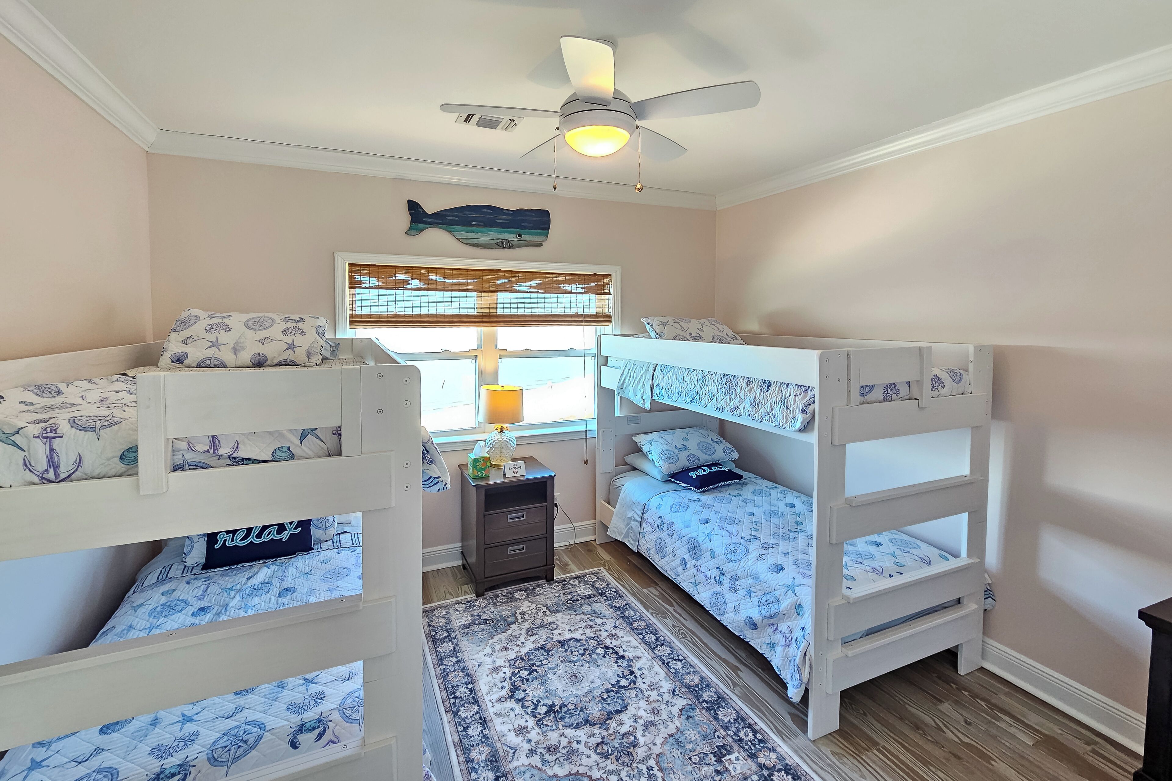 A bedroom with two bunk beds, a ceiling fan, a fish wall decoration, a window with blinds, and a small nightstand with a lamp.