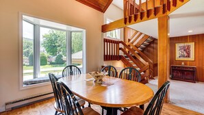 A dining area with a wooden table and chairs, a large window with a view of the outdoors, and a wooden staircase.