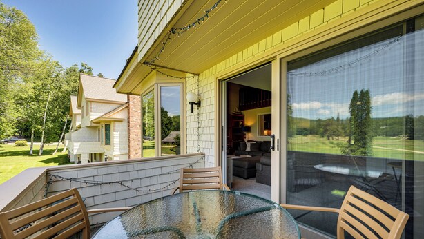 A patio with a glass table and wooden chairs, a brick wall, and a view of a house and trees.