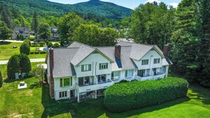 A large, two-story house with a balcony, surrounded by lush greenery and a mountain in the background.