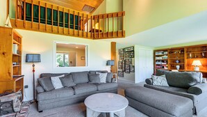 A spacious living room with a high ceiling, a built-in wooden staircase, and a large collection of books and decorative items.