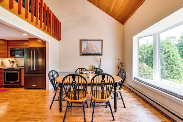 A dining area with a wooden table and chairs, a painting above the table, and a view of trees through large windows. - Beautiful Wentworth Golf Club Condo: Balcony & A/C (Jackson)