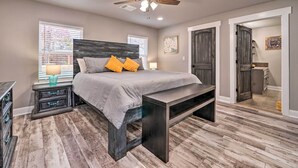 A bedroom with a bed, nightstands, a bench, a ceiling fan, and a door leading to another room. - Lakefront Fort Towson Home: Private Dock & 3 Decks (Fort Towson)