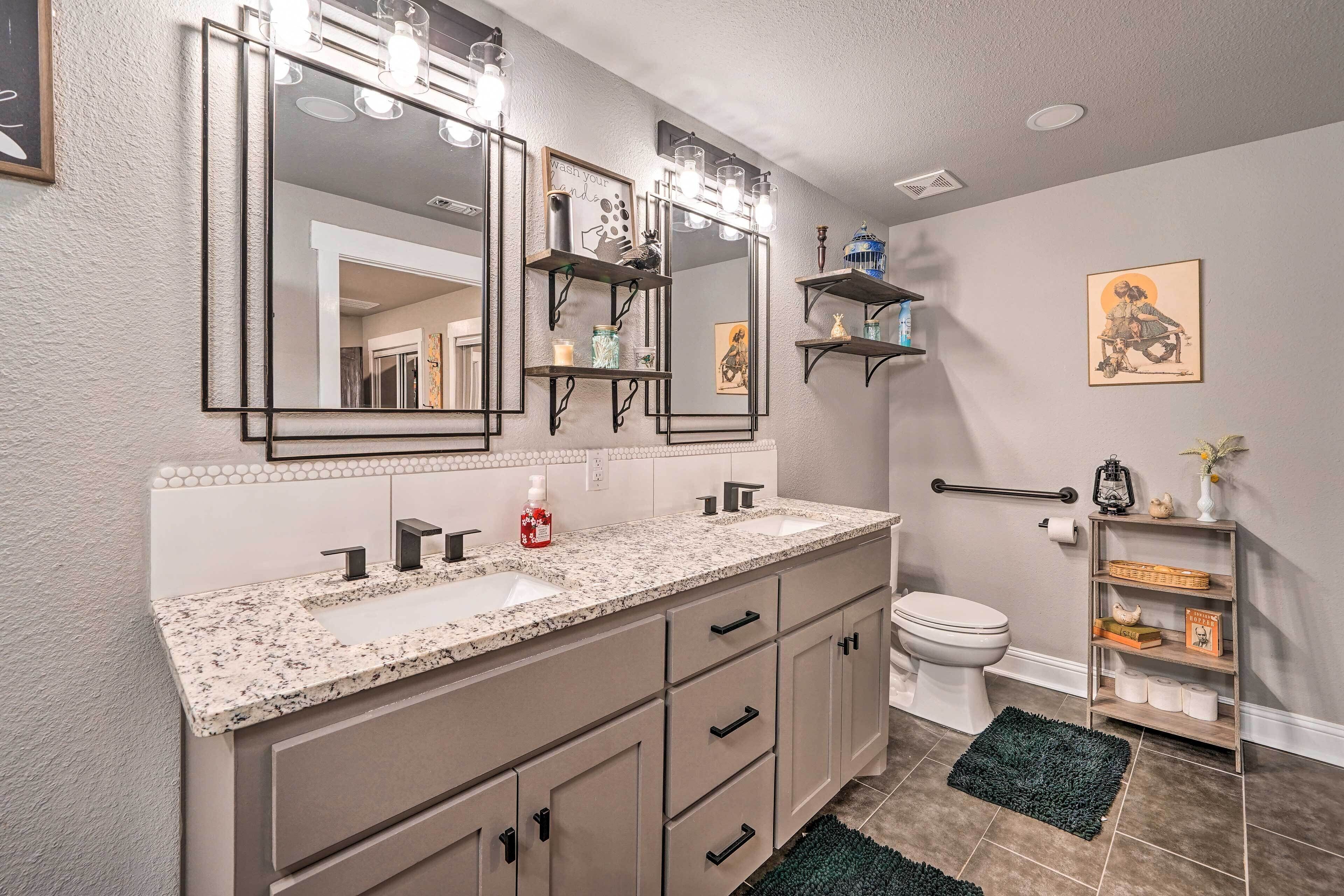 A bathroom with a granite countertop sink, two mirrors, a vanity with drawers, a toilet, and a shelf with toiletries.