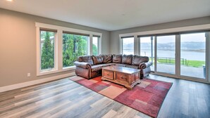 A spacious living room with a large sectional sofa, a wooden coffee table, and a red and beige rug. Large windows provide a view of a lake and greenery. - Scenic & Spacious Getaway on North Bay w/ Deck! (Allyn)