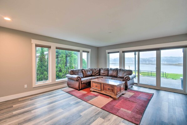 A spacious living room with a large sectional sofa, a wooden coffee table, and a red and beige rug. Large windows provide a view of a lake and greenery. - Scenic & Spacious Getaway on North Bay w/ Deck! (Allyn)