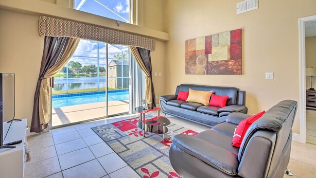 A spacious living room with a large window, a pool view, a leather sofa, and a modern art piece on the wall. - Lake-view Home w/ Game Room in Orlando! (Kissimmee)