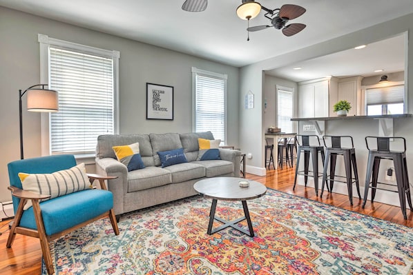 Una sala moderna con sofá, sillón y mesa de centro, una zona de cocina con taburetes de bar y ventanas grandes con persianas.