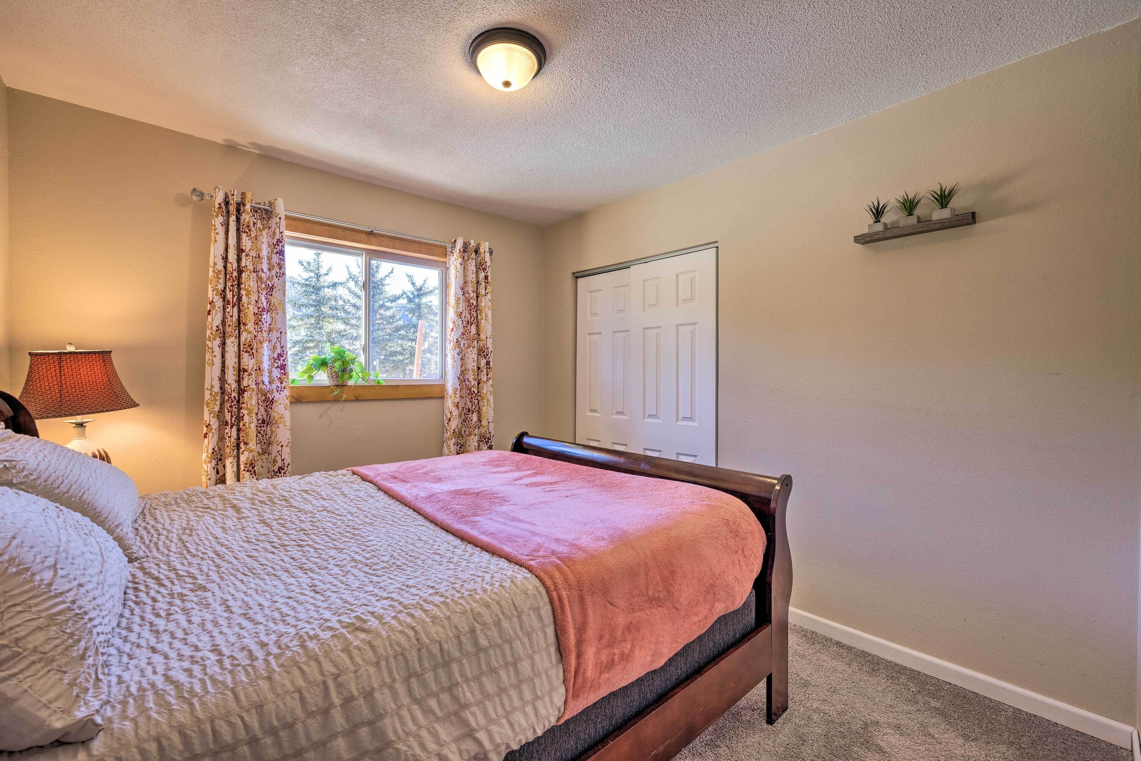 Un dormitorio con cama, ventana con cortinas, lámpara, puerta y estante con plantas.