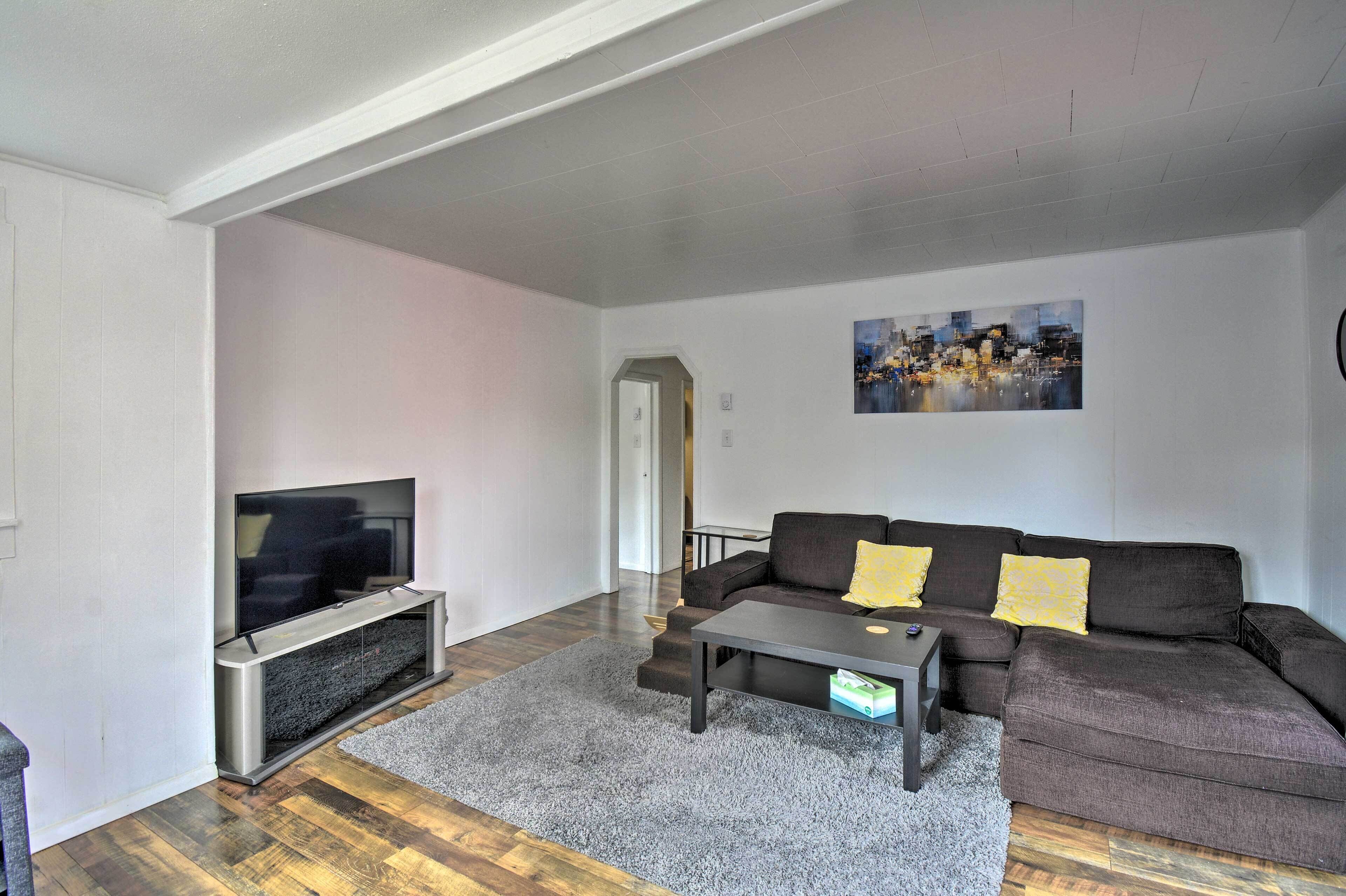 Ein Wohnzimmer mit einem Flachbildfernseher, einem dunkelbraunen Sofa mit gelben Kissen, einem grauen Teppich und einem Stadtbild an der Wand.