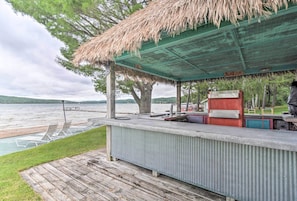 Un espace bar extérieur abrité, doté d’un toit de chaume, un salon au bord de la piscine avec des fauteuils, et une vue sur un lac.