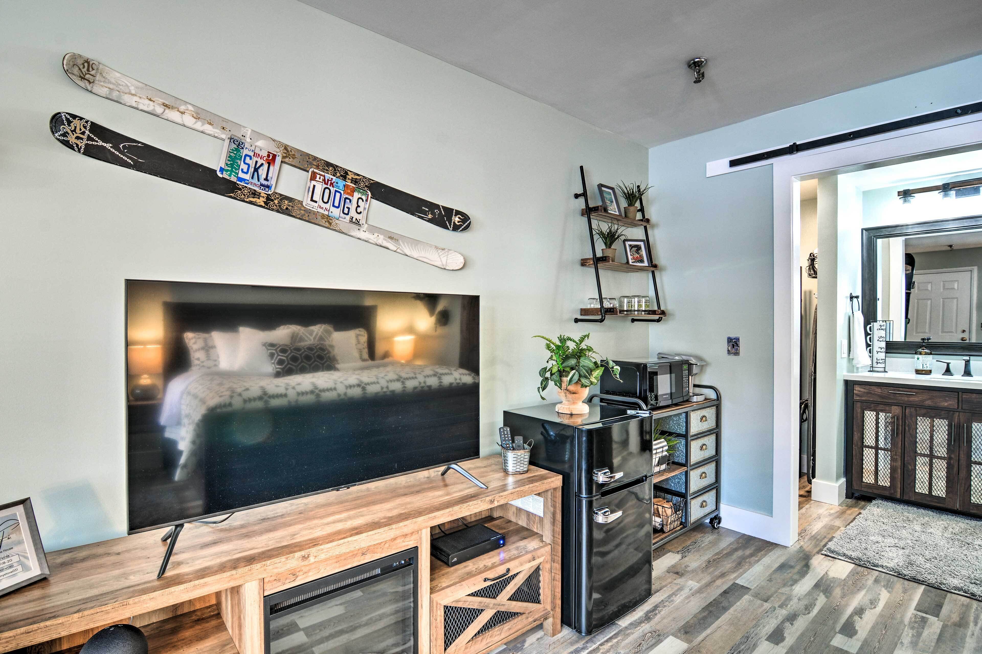 A modern living room with a large flat-screen TV, a wooden entertainment unit, a black refrigerator, and a kitchen area visible through an open doorway.