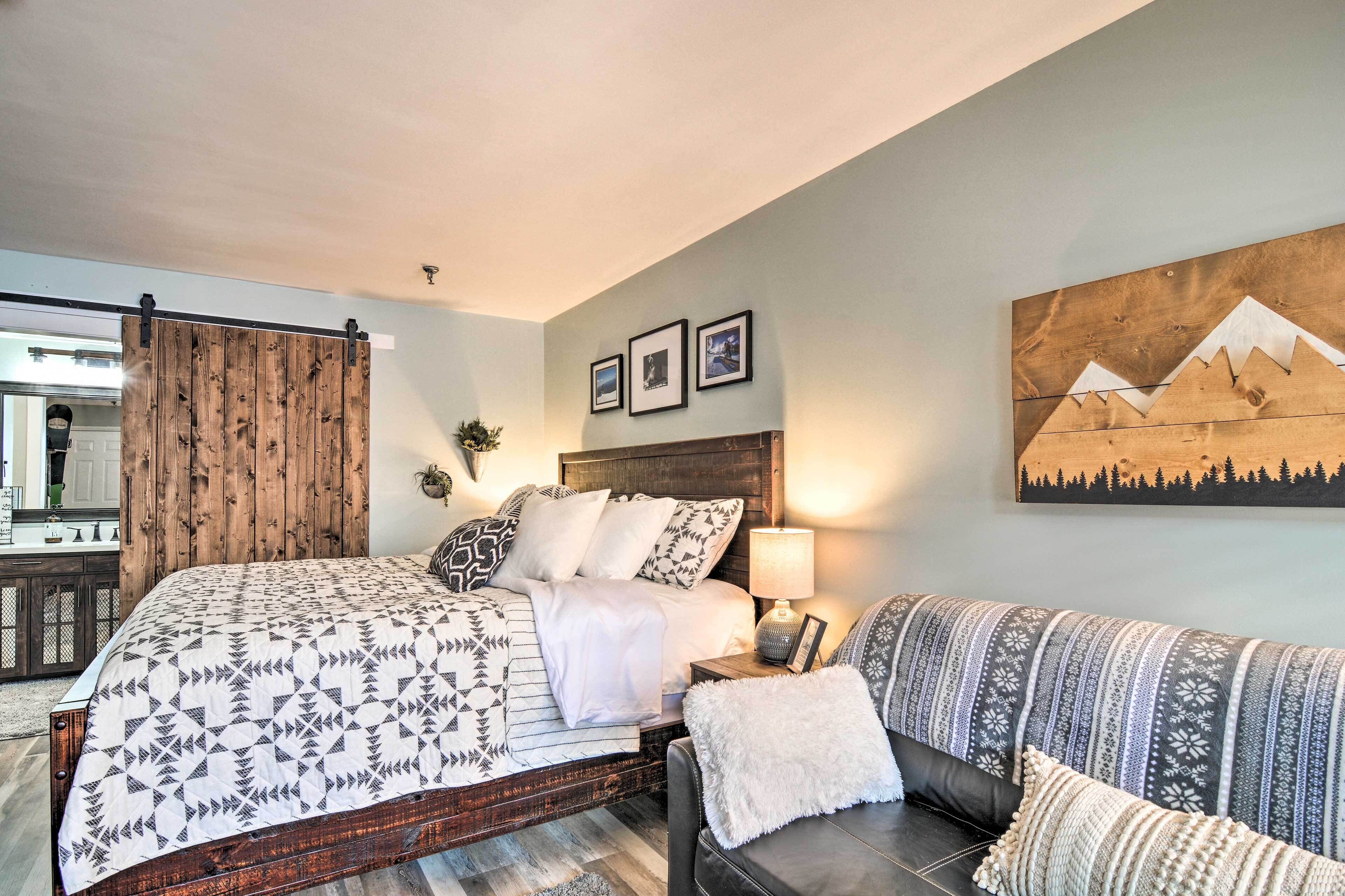 Un dormitorio con cama, mesita de noche, lámpara, sofá y una obra de arte colgada en la pared.
