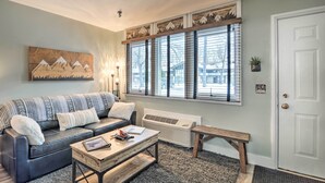 A living room with a sofa, coffee table, and a bench. There is a window with blinds, a door, and a wall-mounted air conditioner. - Ski-in/ski-out Getaway at Boyne Mountain Resort (Boyne Falls)