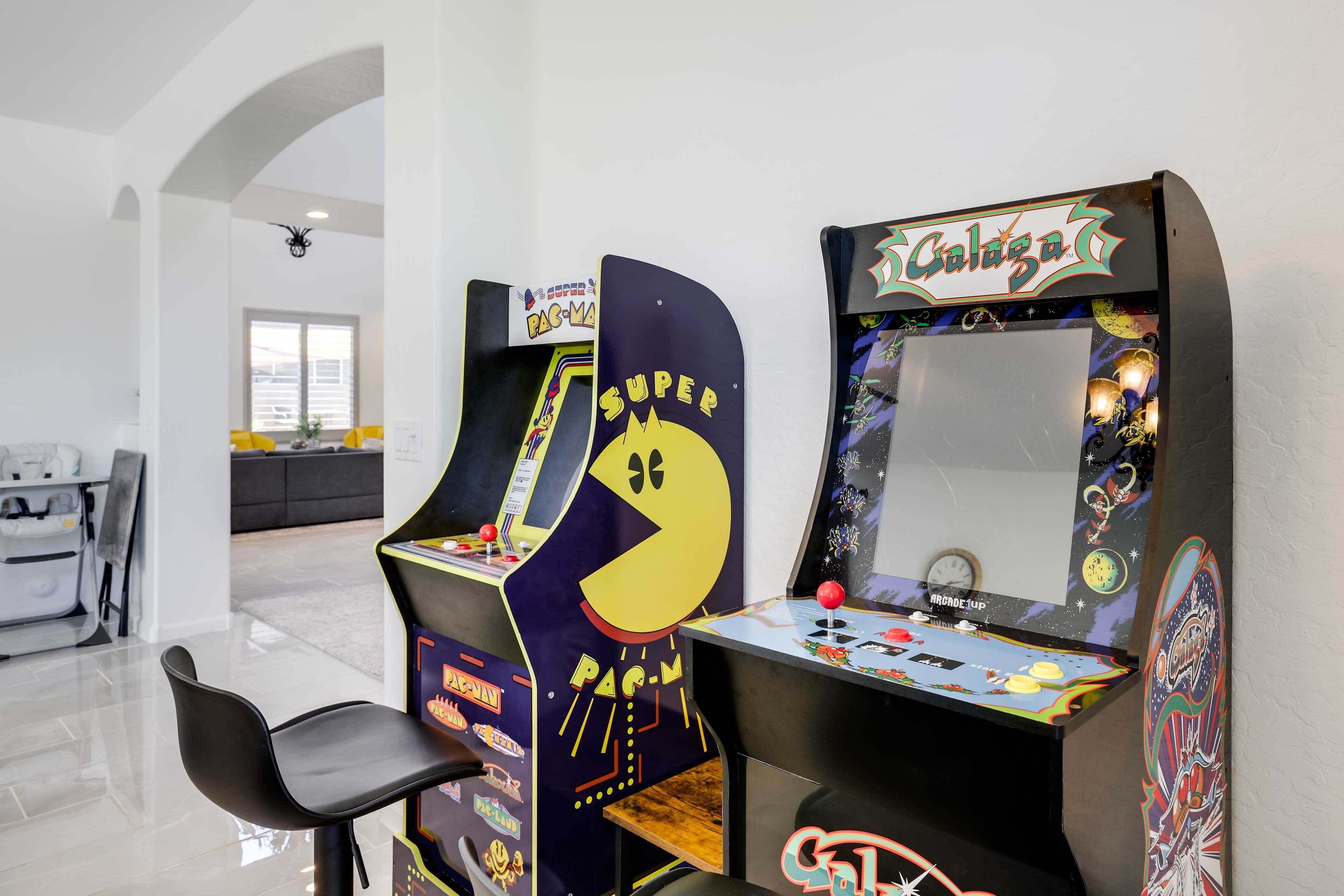 Duas máquinas de arcade com desenhos de Pac-Man e Galaga, uma cadeira preta e um vislumbre de uma área de estar moderna com um sofá e uma mesa de jantar.