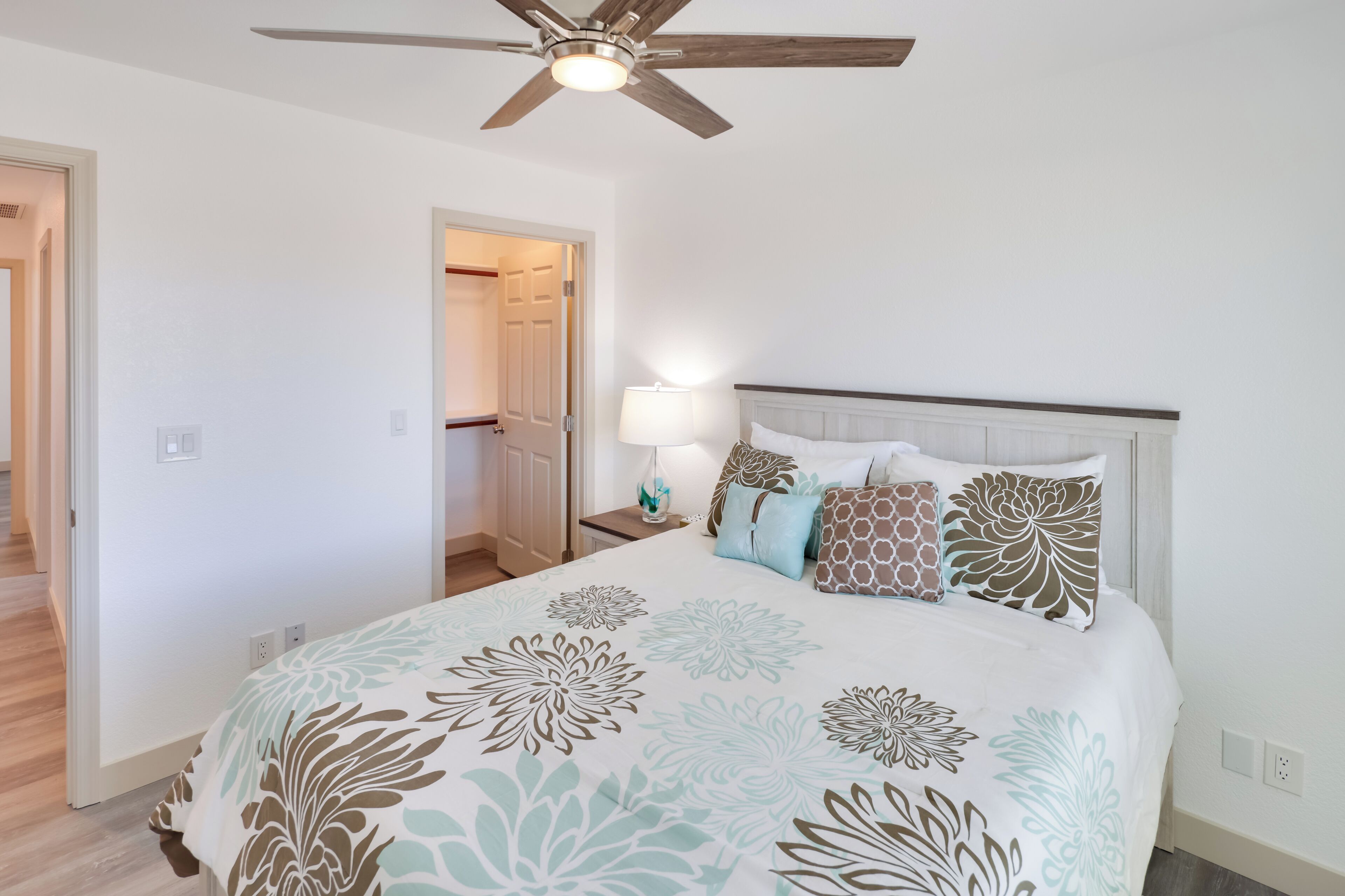 A bedroom with a bed, a ceiling fan, a nightstand with a lamp, and a door leading to another room.
