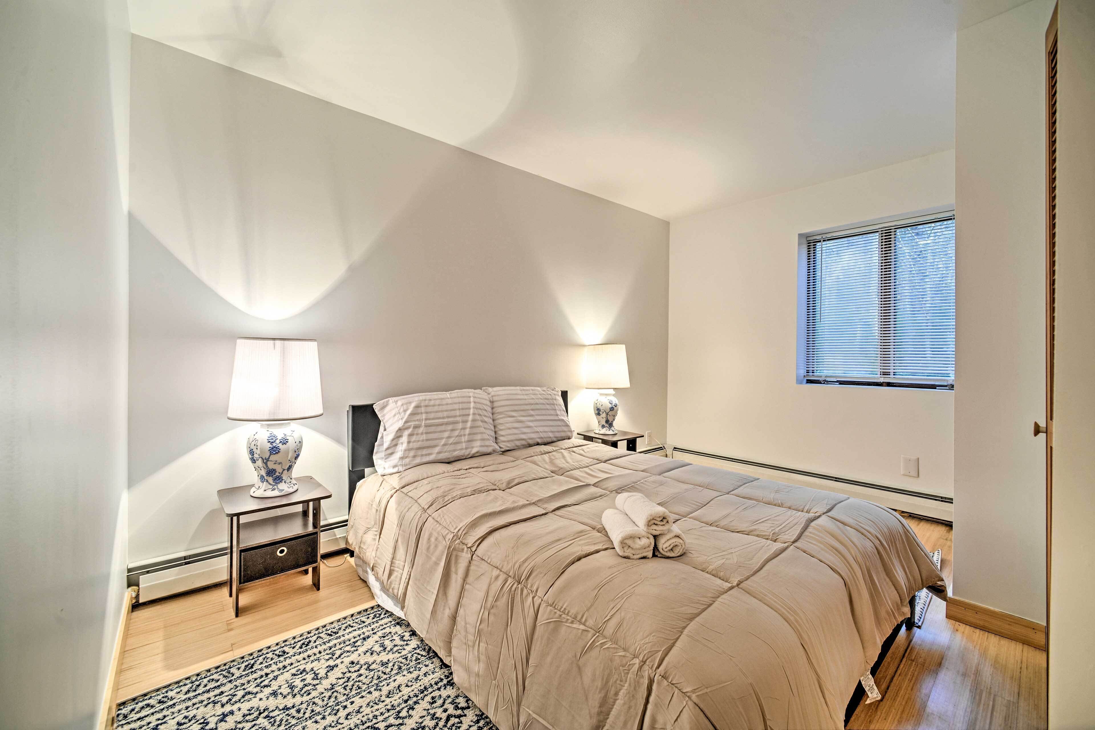 A bedroom with a large bed, two bedside tables with lamps, a window with blinds, and a patterned rug.