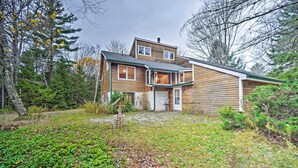 A two-story wooden house with a stone pathway leading to the front door, surrounded by trees and greenery.