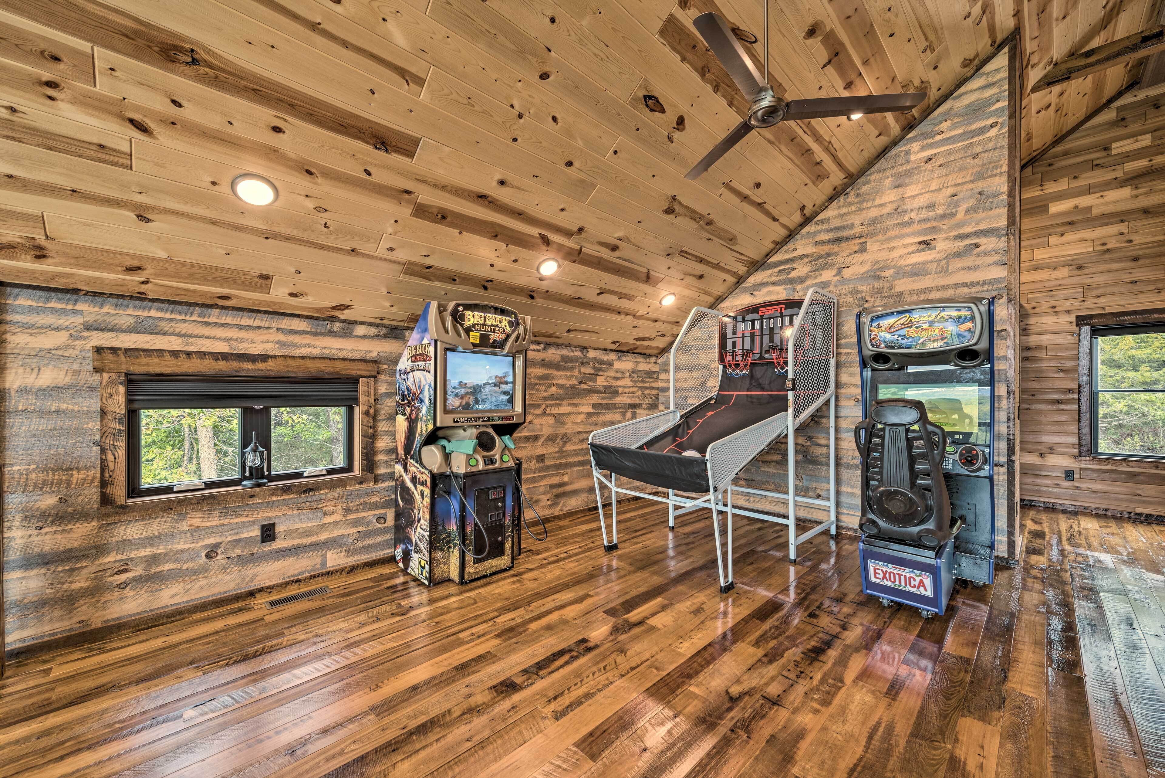 A wooden cabin interior with two arcade games, a bed, and a window overlooking trees.