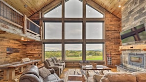 A spacious living room with a large window, wooden ceiling, and a stone fireplace. - Galena Cabin w/ Wraparound Porch & Game Room! (Galena)