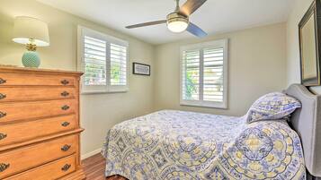 Un dormitorio con cama, cómoda, ventilador de techo y dos ventanas con persianas.