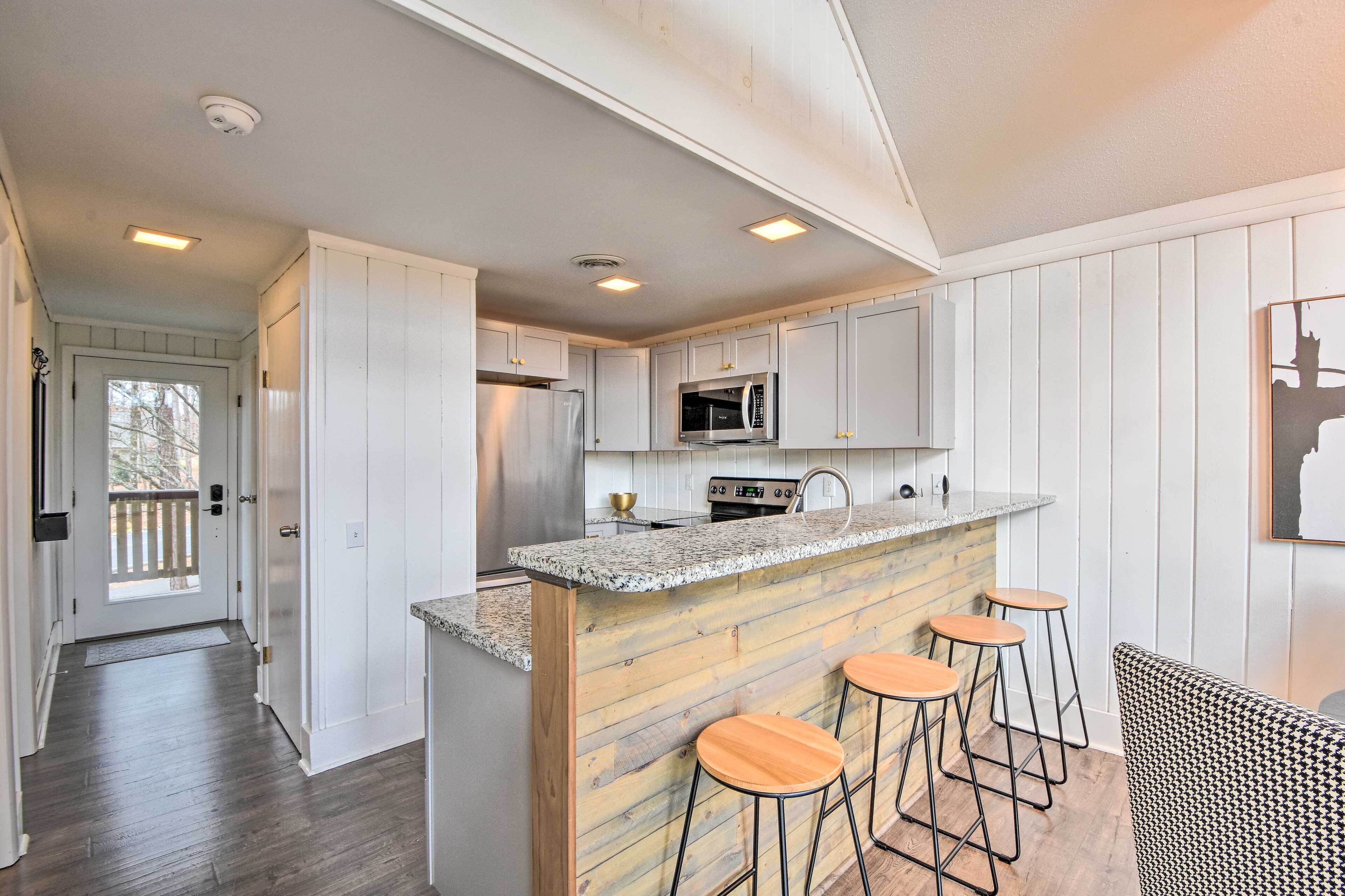A modern kitchen with white cabinets, a granite countertop, and wooden accents. There are two bar stools and a stainless steel sink.