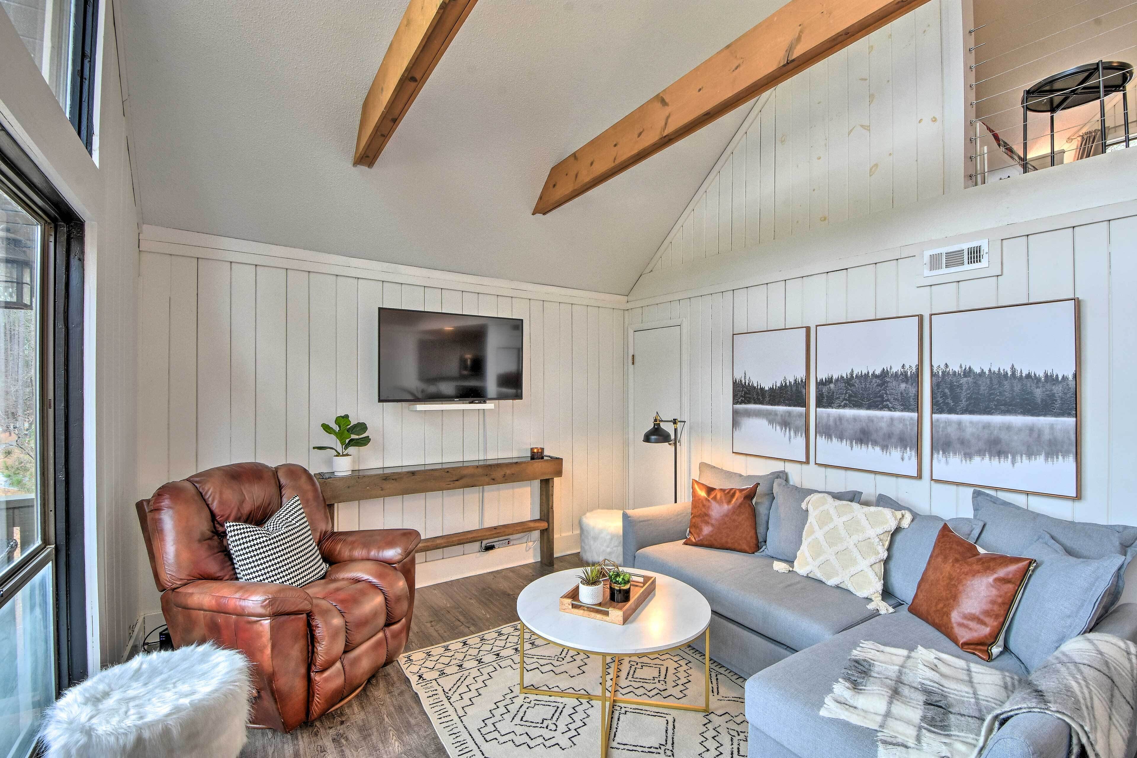 Ein gemütliches Wohnzimmer mit Ledersessel, grauem Sofa und Couchtisch. Ein Fernseher ist an der Wand montiert und ein Fenster lässt natürliches Licht herein.