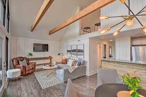 A spacious living area with a high ceiling, wooden beams, a grey sofa, a brown leather chair, and a dining table with a plant.