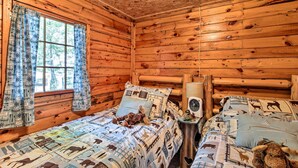 A cozy cabin room with wooden walls, a bed with patterned bedding, a small table with a fan, and a window with curtains. - Rustic Hayward Cabin With Spider Lake Access! (Hayward)
