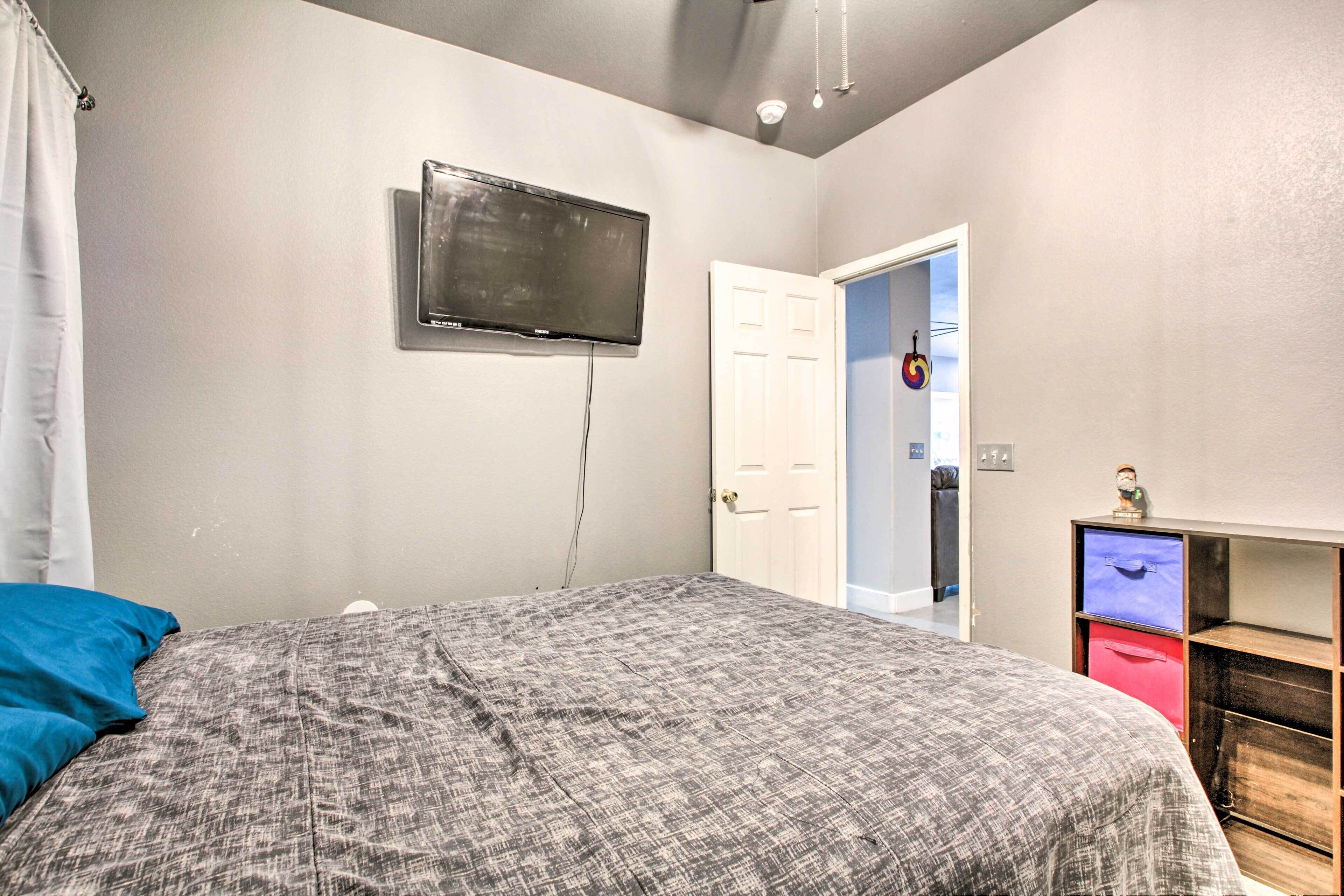 A bedroom with a bed, a television mounted on the wall, a door, and a small shelf with a clock.