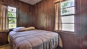 Ein Schlafzimmer mit Holzwandverkleidung, einem Bett mit beige farbenem Überwurf und zwei Fenstern mit Vorhängen.