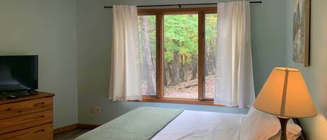 Une chambre à coucher avec un lit, un téléviseur posé sur une commode, une fenêtre avec des rideaux et une lampe.