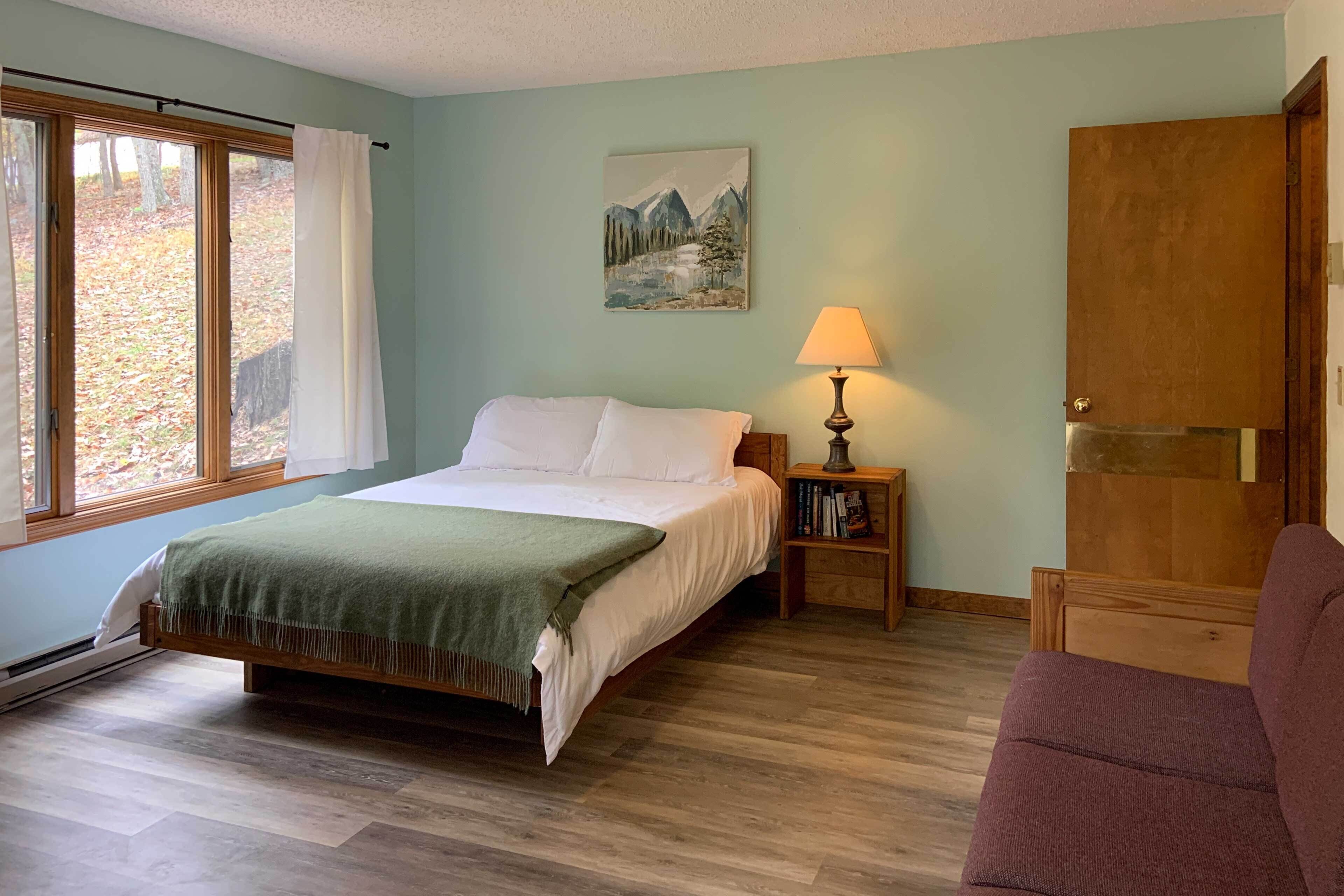A bedroom with a bed, a nightstand with a lamp, a wooden door, and a window with curtains.
