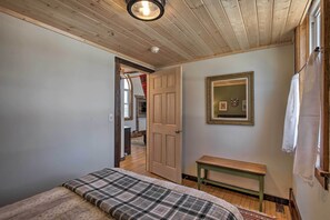 Une chambre avec un plafond en bois, un lit recouvert d’une couverture à carreaux, un banc en bois, un miroir et une porte donnant sur une autre pièce.