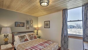 A bedroom with a wooden ceiling, a bed with a plaid blanket, a nightstand with a lamp, and a window with curtains. - Renovated Historic Church Along Namekagon River! (Hayward)