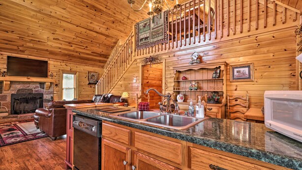 A wooden cabin interior with a stone fireplace, a kitchen with granite countertops, and a staircase leading to an upper level. - Douglas Lake Cabin w/ Boat Ramp & Water Access! (Sevierville)