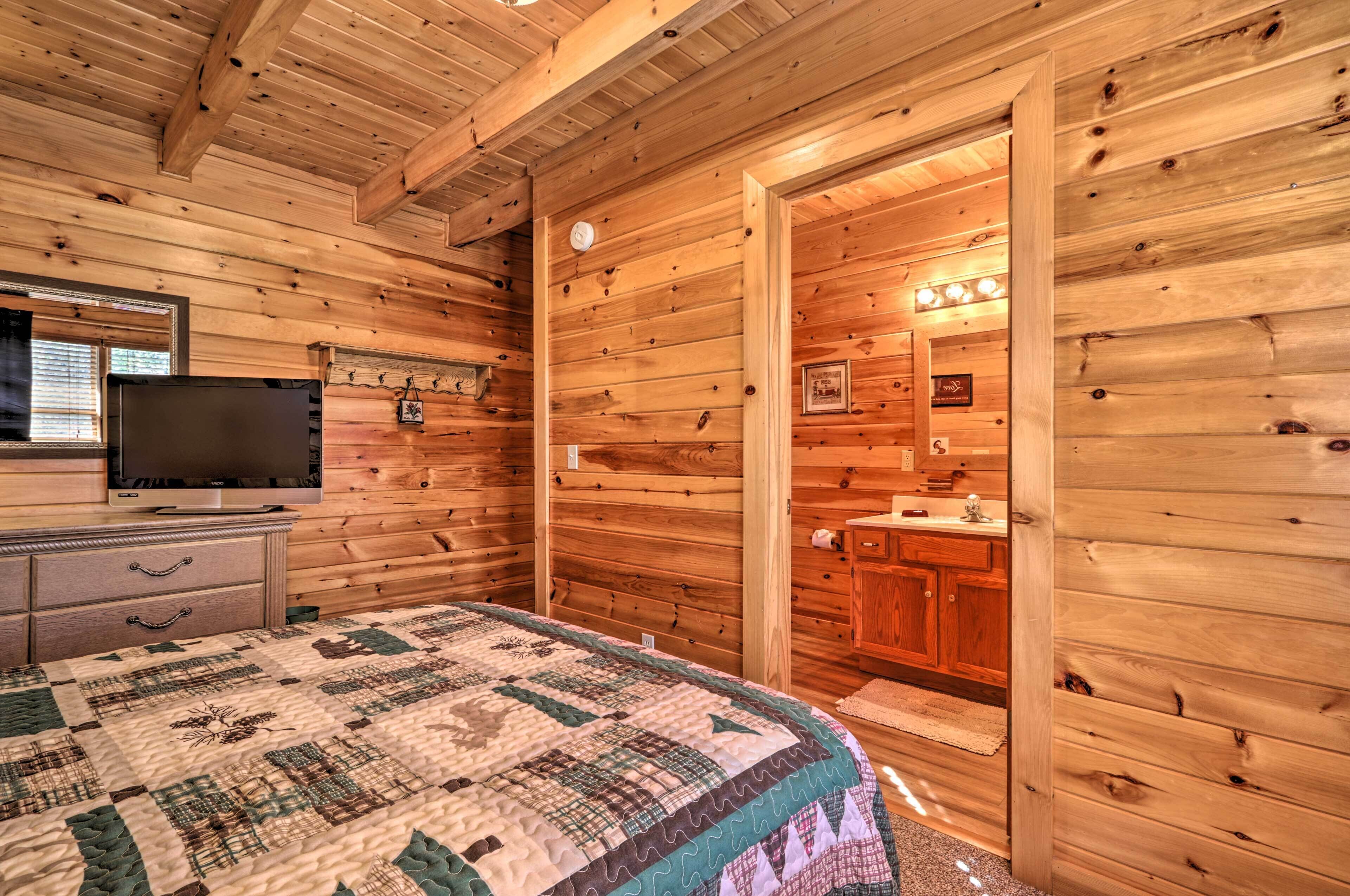 Een slaapkamer in een houten hutje, met een bed, een dressoir, een televisie en een zichtbare badkamer door een geopende deur.