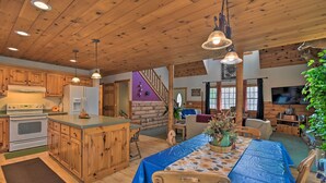 A rustic cabin interior with wooden ceilings, a central island with a sink, a dining table with blue tablecloth, and a fireplace.