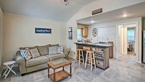 A living room with a sofa, coffee table, and a small side table. There is a kitchen area with stools and a countertop.