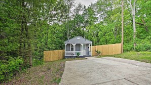 A small, single-story wooden structure with a porch, surrounded by dense green trees and a wooden fence.