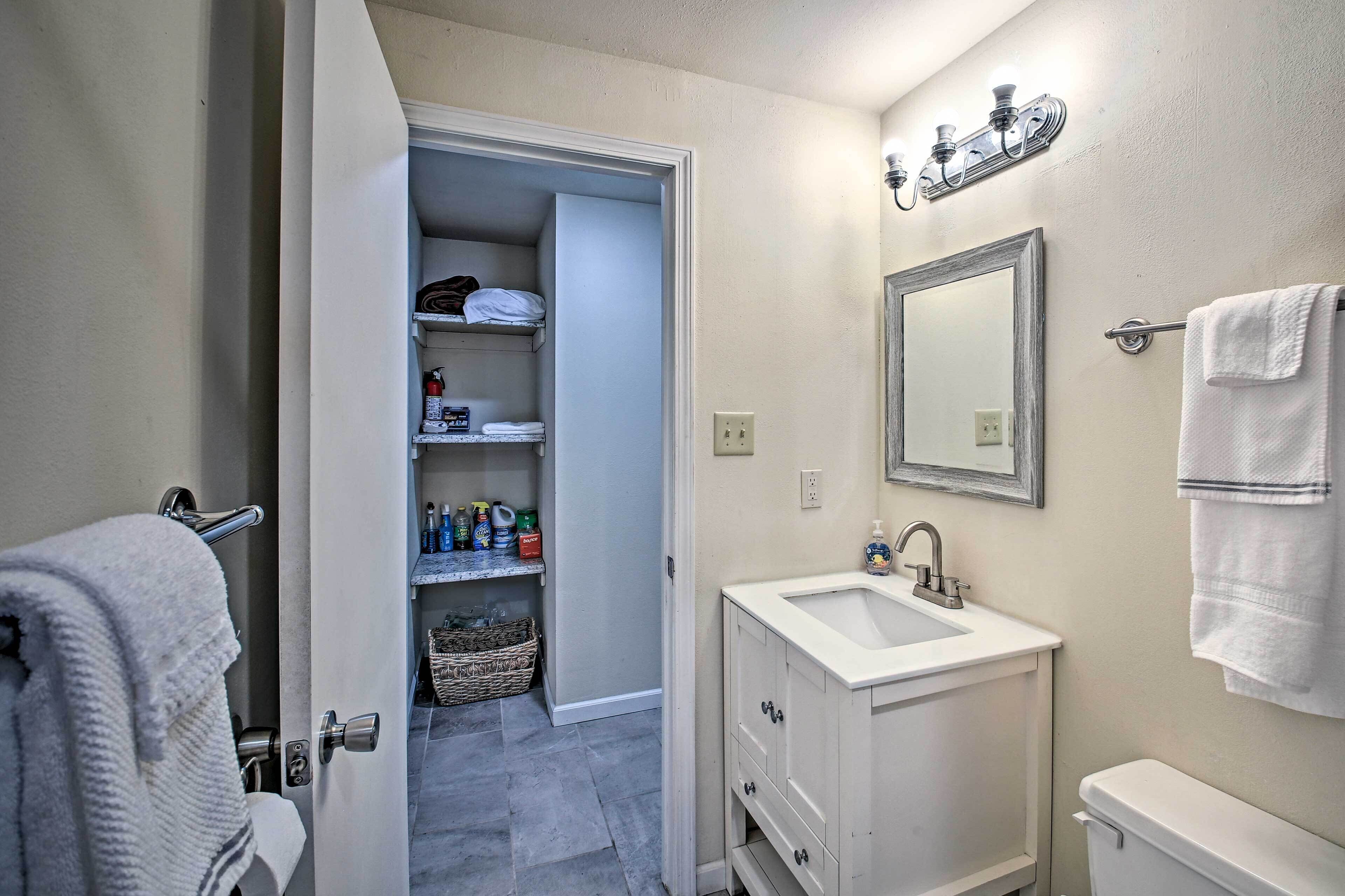 Un baño con lavamanos blanco, espejo y toallero. Se ve una puerta que da a un armario con repisas y cestas.