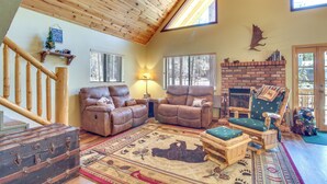 A cozy living room with a fireplace, leather sofas, a wooden floor, and a large window. - 9 Mi to Rainbow Lake: Woodsy Pinetop Cabin w/ Deck (Pinetop)