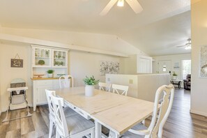 Uma área de refeições com mesa de madeira, cadeiras brancas, um armário branco com portas de vidro e um ventilador de teto.